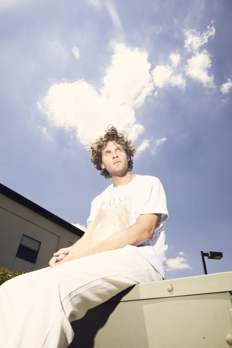 joe p sits on a ledge, wearing an all white outfit with the blue, clouded sky behind him.