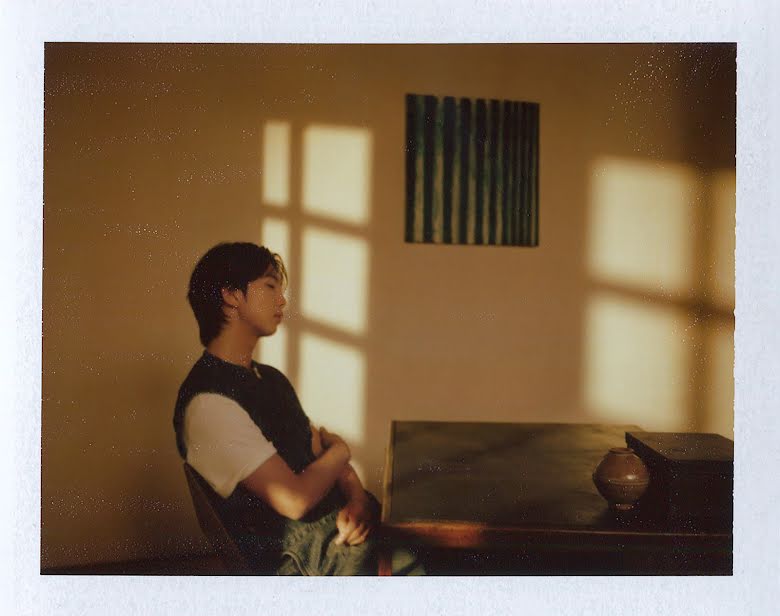Promotional image for RM's album Indigo. RM sits leaned back at a table. A work by Korean Painter Yun Hyong-keun hangs on a white wall behind him. Sun streams diagonally into the room from out-of-frame windows.